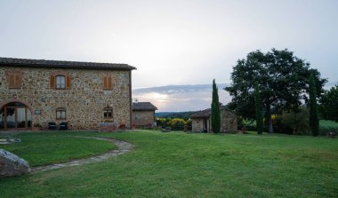 İtalya 'nın Pienza kenti yakınlarındaki Toskana kırsalında yeşil çimenler, uzun selvi ağaçları ve terasotta saksılarıyla çevrili geleneksel taş çiftlik evi, gün batımında sakin bir bahar akşamı yakalandı..