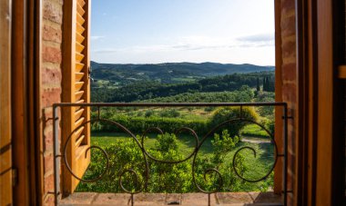 View through a rustic wooden window reveals rolling Tuscan hills with olive groves, cypress trees, and lush vegetation. Wrought iron balcony railing frames the picturesque countryside scene.