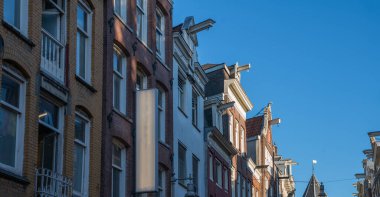 Historic gabled facades with traditional hoisting beams and tall windows in the old center of Amsterdam, Netherlands. The narrow buildings are bathed in evening sunlight under a clear blue sky.
