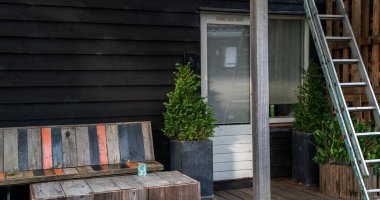 Rustic outdoor space with a weathered wooden bench featuring colorful painted slats, a potted evergreen shrub, a metal ladder leaning on a black wooden wall, and a window with a shade.