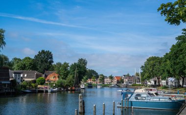 Blokzijl, Overijssel köyündeki kanal, demirlemiş tekneler, geleneksel evler ve açık bir yaz gökyüzünün altında yeşil ağaçlar ve huzurlu binalarla çevrili beyaz bir asma köprü ile dolu.