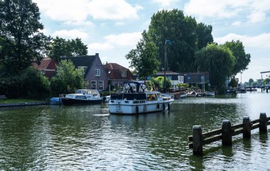 Hollanda 'nın Weesp kentindeki kanalda yol alan tekne, mavi gökyüzü altında demirli gemiler, tuğlalı evler ve yeşil ağaçların arasından geçiyor, asma köprüler ve sessiz su yolu boyunca görünen uzak bir yel değirmeni bulunuyor..