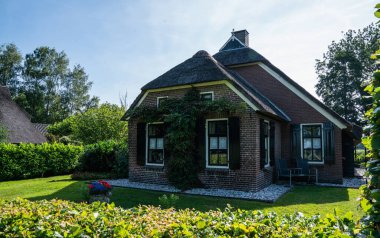 Yazın Hollanda 'nın kırsal kesimlerinde çeşitli yaprak döken ağaçlarla çevrili sık yeşil çalıların ve çiçek açan çiçeklerin arkasına kısmen saklanmış çatı evi..