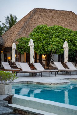 Lüks tropikal tatil köyü, sazdan çatılı bir bar, beyaz çiçekli yemyeşil ağaç, minderli ahşap şezlonglar, ve çevreyi yansıtan turkuaz yüzme havuzu.