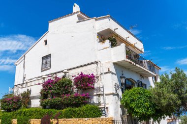 Canlı bougainvillea ile süslenmiş beyaz İspanyol tarzı bir bina. Arka planda demir parmaklıklı balkonlar ve mavi bir gökyüzü var..