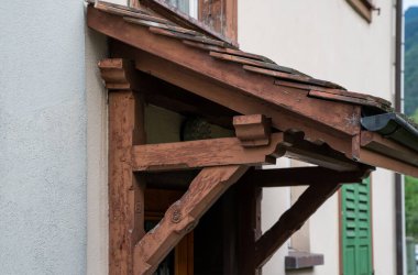 Wooden porch canopy with decorative corbels, angled supports attached to a cream plastered building in a European village, featuring aged beams, clay roof tiles, and a green shutter in the background.