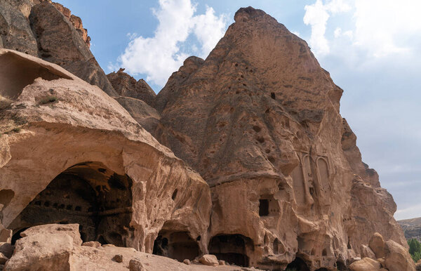 an impressive rock formation pocked with numerous cave openings, ancient dwellings carved into a rugged cliff face. The cloudy sky above