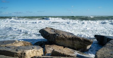 Ön planında büyük düz kayalar olan kıyı deniz manzarası ve dalgalı deniz dalgaları sahile çarpıyor. Gökyüzü bulutlarla temizlendi. Parlak güneş ışığı, ıslak taşın dokusunu vurgular.