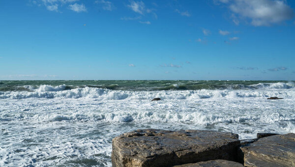 Прибрежный морской пейзаж с большими плоскими скалами на переднем плане и турбулентными морскими волнами, падающими на берег. Небо чистое с рассеянными облаками. Яркий солнечный свет подчеркивает текстуру влажного камня.