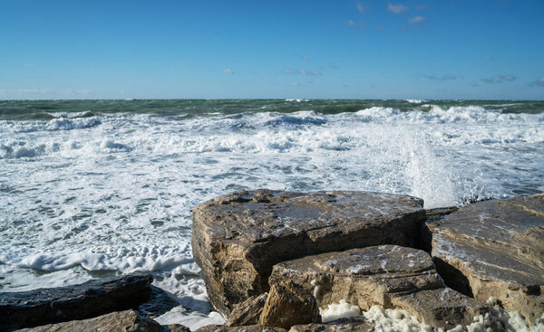 Прибрежный морской пейзаж с большими плоскими скалами на переднем плане и турбулентными морскими волнами, падающими на берег. Небо чистое с рассеянными облаками. Яркий солнечный свет подчеркивает текстуру влажного камня.