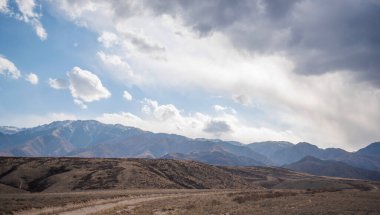 Gökyüzü engebeli sıradağların üzerine gölgeler düşürüyor, tepeler karla kaplanıyor. Dağ etekleri, seyrek bitki örtüsü ve dolambaçlı bir yol ile dinamik bir gökyüzünün altındaki görkemli dağlara doğru yol alır.