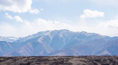 berrak mavi gökyüzünün altındaki dağlık arazi. Kumral renklerle kaplanmış yuvarlanan tepeler, uzaktaki engebeli, karla kaplı tepelerle tezat oluşturuyor. Dağınık bulutlar gökyüzünü süslüyor