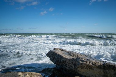Ön planında büyük düz kayalar olan kıyı deniz manzarası ve dalgalı deniz dalgaları sahile çarpıyor. Gökyüzü bulutlarla temizlendi. Parlak güneş ışığı, ıslak taşın dokusunu vurgular.