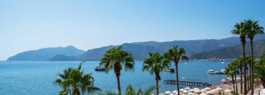 Huzurlu bir sahil sahnesi. Güneşli şezlonglar ve sakin mavi denize bakan şemsiyeler. Palmiye ağaçları kumlu sahili çerçevelerken, tekne ve su aktiviteleriyle dolu bir rıhtım suya doğru uzanıyor..