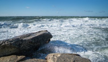 Ön planında büyük düz kayalar olan kıyı deniz manzarası ve dalgalı deniz dalgaları sahile çarpıyor. Gökyüzü bulutlarla temizlendi. Parlak güneş ışığı, ıslak taşın dokusunu vurgular.