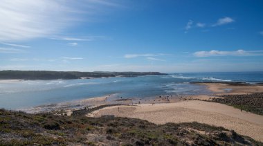 Kumlu plajı ve kıyı boyunca kayalık oluşumları olan çarpıcı bir kıyı manzarası. Sakin okyanus, berrak mavi gökyüzünün altında ufka kadar uzanıyor..