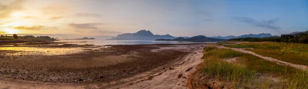 Amnesvika körfezindeki batan güneşin ufukta görünen panoramik manzarası. Kumsal, patikanın etrafındaki çimenler. Ada Amnoya 'da günbatımı. Yazın Norveç