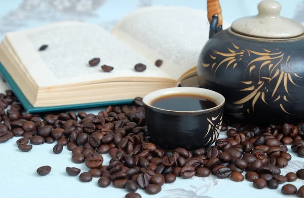 Tasse Kaffee, Kaffeebohnen, eine Teekanne im Hintergrund ein geschlossenes Buch — Stockfoto