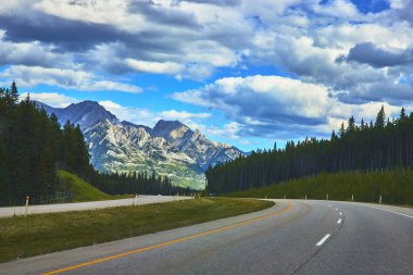 Banff Canada 'da yüksek dağlar ve dramatik bulutlarla dolu yoğun bir orman boyunca uzanan manzaralı bir otoyol seyahat ve macera için ilham verici bir sahne oluşturuyor..
