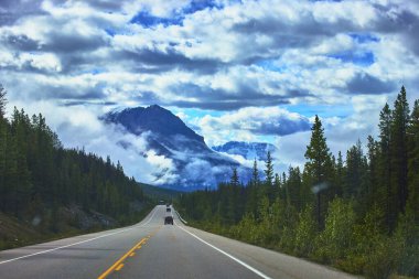 Jasper Kanada 'da manzaralı bir otoyol bereketli bir çam ormanında dramatik bulutlar ve yükselen görkemli bir dağ zirvesi boyunca esiyor. Bu ikonik seyahat yolunun vahşi güzelliğini yakalıyor..
