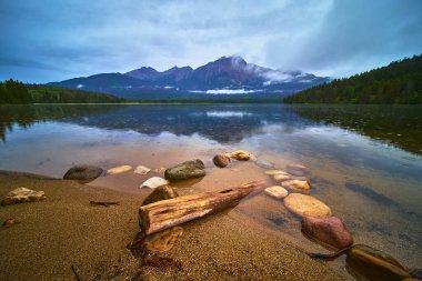 Jasper Kanada 'daki Piramit Gölü' nün sakin manzarası. Durgun su dağları ve ormanı yansıtıyor kasvetli gökyüzünün altında kumlu kıyılar ve dağınık taşlar var..