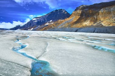 Jasper Canada 'daki Athabasca Buzulu' nun yüzeyinden geçen parlak mavi erimiş su akıntıları arka planda yükselen engebeli dağlar ve yukarıda canlı bir gökyüzü.