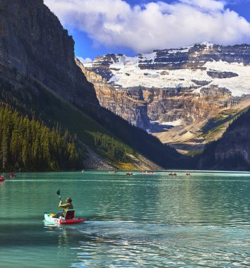 Bir insan Banff 'taki Louise Gölü' nün turkuaz sularında kanolar sürüklenirken ve el değmemiş gölün üzerinde yükselen buzul kaplı bir dağ sonsuz yeşil ormanla çevriliyken kano kürek çeker..