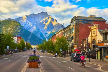 Kanada 'daki Banff Kasabası' nın canlı ana caddesinin arkasında yükselen güneş ışığı, bisikletçiler ve çiçek açan insanlarla dolu, canlı ve hoş bir yaz ortamı yaratıyor..