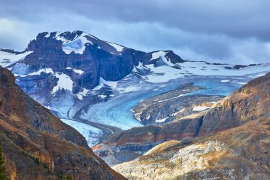 Banff Canada 'da kasvetli bir gökyüzünün altındaki kayalık dağların arasında dramatik bir buzulun akışı buzul kaplı zirvelerin ve engebeli yaban arazilerin saf güzelliğini yakalıyor..