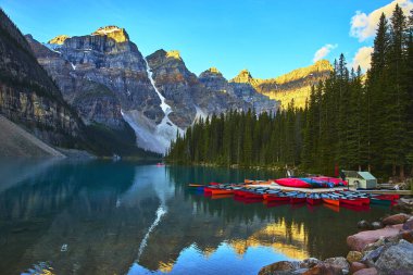 Renkli kanolar, Banff Canada 'daki Moraine Gölü' nün berrak sularında dinlenir. Gün doğumunda altın saat güneş ışığı yükselen dağları aydınlatır ve çam ağaçlarını yansıtır..