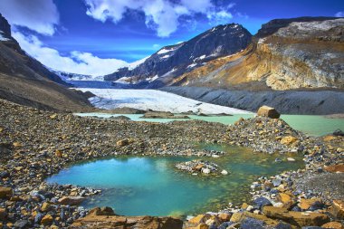 Canlı turkuvaz eriyen su havuzu Jasper Kanada 'daki Athabasca Buzulu' nun altındaki kayalık arazide bulunur. Dramatik dağ zirveleri ve parlak bir yaz gökyüzü nefes kesici bir doğal manzara yaratır..