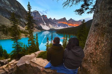 İki turist, Banff Canada 'daki Moraine Gölü' nün üzerinde nefes kesici gündoğumunun keyfini çıkarıyorlar. Canlı mavi suları ve dramatik dağ manzaralarıyla bir merak ve macera hissi yaratıyorlar..