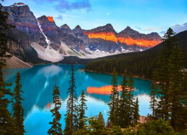 Moraine Gölü 'nün yukarısındaki Banffs dağlarında gün doğumu ışığı parıldıyor. Altın saat renkleri yoğun yeşil ormanlarla çevrili berrak turkuaz suları yansıtıyor. Huzurlu ve ilham verici bir manzara yaratıyor..
