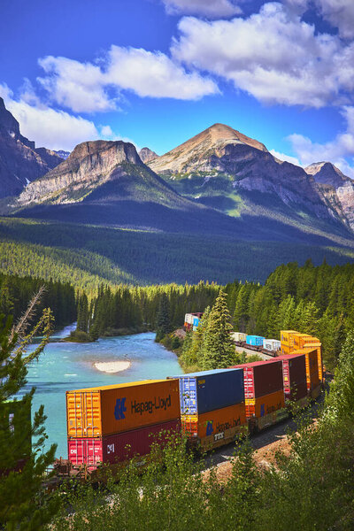 A freight train rounds Morants Curve in Banff Canada with vibrant shipping containers following a turquoise river below towering mountain peaks under a bright sky.