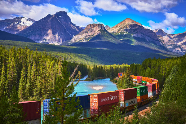 A freight train navigates Morants Curve in Banff Canada surrounded by evergreen forests and towering mountains with a vibrant blue river flowing alongside for a stunning scenic view.