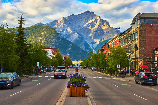 Colorful flowers in a street planter brighten the lively main avenue of the Town of Banff Canada while majestic mountains rise in the background under a vibrant summer sky.