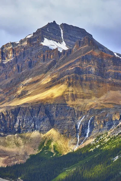 Güneş ışığı, Banff ve Peyto Gölü yakınlarındaki Kanada Kayalıkları 'ndaki yoğun yeşil bir ormanda şelalenin çağlayarak yükseldiği engebeli bir dağ zirvesini vurguluyor..