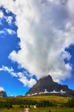 Yürüyüşçüler, Buzul Ulusal Parkı Montana 'daki Saklı Göl Yolu' ndaki dramatik bulutların altında yükselen bir dağ zirvesine doğru, yemyeşil çayırlar boyunca manzaralı bir patikayı takip ediyor..