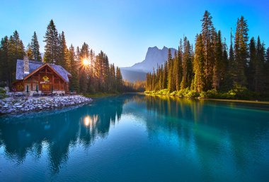 Altın saat güneş ışığı, Yoho Kanada 'daki Emerald Gölü' ndeki kır evinin yanındaki ağaçların arasından süzülüyor. Sakin turkuaz suyu ve huzurlu manzarayı tamamlayan yüksek bir dağ..