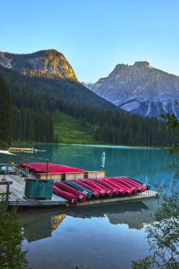 Kırmızı kanolar, Emerald Gölü 'nün turkuaz sularında sessiz bir rıhtım oluşturur. Dağlar arka planda yükselir ve güneş erken doğar. Yoho Kanada' nın huzurlu manzarasını ön plana çıkarır..