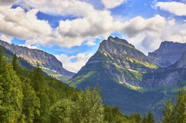 Buzul Ulusal Parkı 'nda parlak bir gökyüzü altında yoğun yeşil ormanların üzerinde yükselen görkemli dağlar, Montana' da vahşi doğayı ve ABD 'nin doğal güzelliklerini yakalıyor..