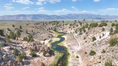 Mammoth Gölleri yakınlarındaki Hot Creek Jeolojik Arazisi 'nin üzerinde uçan kanyon engebeli uçurumları ve açık mavi gökyüzünün altında gün ışığında uzak dağ sıraları yer alır..