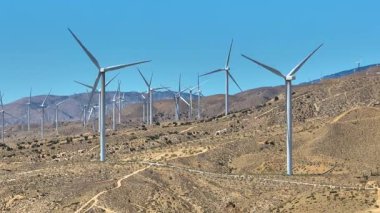Mojave Çölü 'ndeki rüzgâr türbinlerinin üzerinde sabit duran hava görüntüleri kurak tepelere karşı sıralanmış türbinleri yakalıyor ve yenilenebilir enerjiyi gösteren açık mavi bir gökyüzü ve