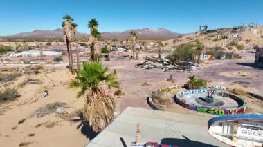 California 'daki terk edilmiş Dolores Gölü Su Parkı' nda uçan hava uçuşu graffiti kaplı harabeleri, palmiye ağaçları ve engin çöl manzaralarını berrak mavi gökyüzünün altında ileri doğru hareket halinde gösteriyor..