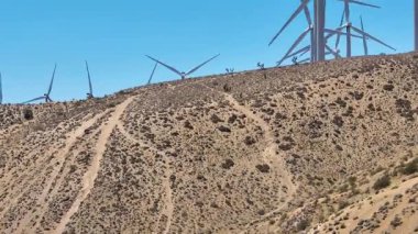 Mojave Çölü tepelerinde, yenilenebilir enerji ve uçsuz bucaksız kurak araziyi vurgulayan, berrak Kaliforniya gökyüzünün altındaki rüzgar türbinlerini ve engebeli patikaları gözler önüne seren hava kaidesi görüntüsü yükseliyor..