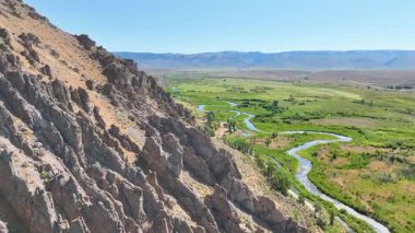 Engebeli dağ uçurumlarının üzerinden ve Legion Park Cokeville Wyoming 'de dolambaçlı bir nehirden uçan bir hava aracı. Kamera ileri doğru süzülüyor. Geniş vadi manzaraları ve yemyeşil yeşil bir alan.