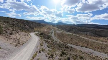 Las Vegas ve Charleston Dağı yakınlarındaki güneşli Nevada çölünde uçan FPV virajlı toprak yollar engebeli arazi ve bulutlarla kaplı gökyüzü altındaki dramatik dağ manzarası.