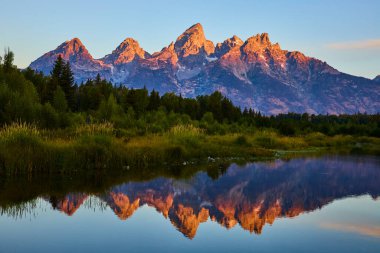 Gün doğumu ışığı, Wyoming 'deki Schwabacher Landing' deki Grand Teton dağlarının tepelerini doldurur. Yansımaları, yemyeşil ve sakin sabahlarla çevrili sakin sularda parıldarken altın bir parıltıyla.