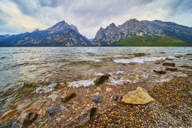 Dalgalar Jenny Gölü 'nün kayalık kıyı şeridine vuruyor. Yükselen Grand Teton dağları berrak suyun ötesine yükseliyor. Huysuz Wyoming gökyüzünün altında..