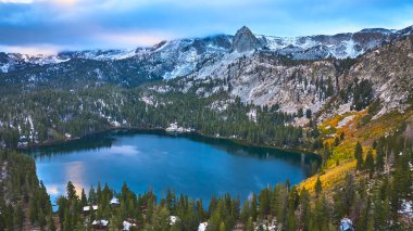 Yoğun çam ormanı ve Mamut California yakınlarında karla kaplı dağlarla çevrili derin mavi bir göl manzarası. Yumuşak ışık, sonbahar renklerine ve dramatik yansıyan sakin suya vurgu yapıyor